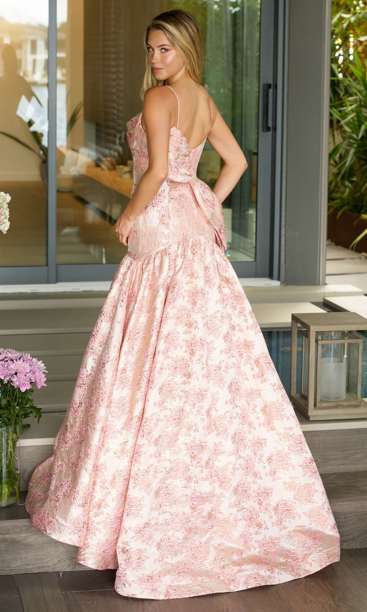 A woman stands on a porch in the Drop-Waist Long Print Prom Gown (Ava Presley 26419), featuring a floral print and back bow, looking over her shoulder at the camera.